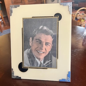 May include: A vintage portrait photo in a cream-colored frame with black and silver accents. The photo features a smiling man in a suit. The frame has decorative corners and black circles. The photo is signed "Ronald Reagan".