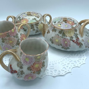 May include: A vintage porcelain tea set featuring a teapot, sugar bowl, and two teacups. The set is decorated with colorful floral and butterfly motifs. The handles and rims have gold accents. The tea set is placed on a white doily.