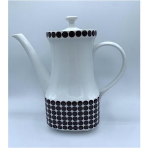 Może przedstawiać: Biały ceramiczny dzbanek do kawy z długim dziobkiem, zakrzywionym uchwytem i pokrywką. Dzbanek jest ozdobiony wzorem czarnych kropek na górze i na dole. Projekt jest klasyczny i minimalistyczny.