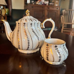 May include: A vintage white porcelain tea set with gold trim and decorative vertical stripes. The set includes a teapot with a lid and a creamer. The teapot has a curved spout and handle, and the creamer has a curved handle.