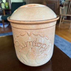 May include: A tan ceramic Rumtopf pot with a matching lid. The pot has a decorative oval design with the word "Rumtopf" in script. The pot is cylindrical with horizontal lines and a textured surface.