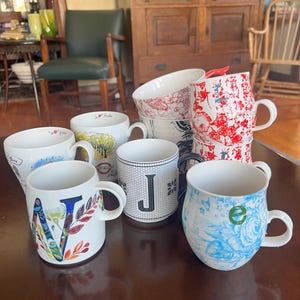 K&ouml;nnte beinhalten: Eine Sammlung von Keramikbechern mit verschiedenen Designs, darunter Blumenmuster und Buchstaben. Die Becher sind wei&szlig; mit farbigen Akzenten und stehen auf einer dunkelbraunen Oberfl&auml;che.