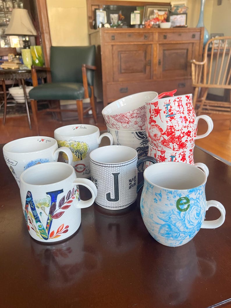 K&ouml;nnte beinhalten: Eine Sammlung von Keramikbechern mit verschiedenen Designs, darunter Blumenmuster und Buchstaben. Die Becher sind wei&szlig; mit farbigen Akzenten und stehen auf einer dunkelbraunen Oberfl&auml;che.