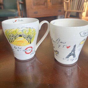 K&ouml;nnte beinhalten: Zwei wei&szlig;e Keramikbecher mit handgezeichneten Illustrationen. Ein Becher zeigt eine Londoner Szene mit dem Text "Hyde Park" und "Padding Hill Gate". Der andere Becher hat ein Pariser Thema mit einer Eiffelturm-Illustration und dem Text "Paris je t'aime".