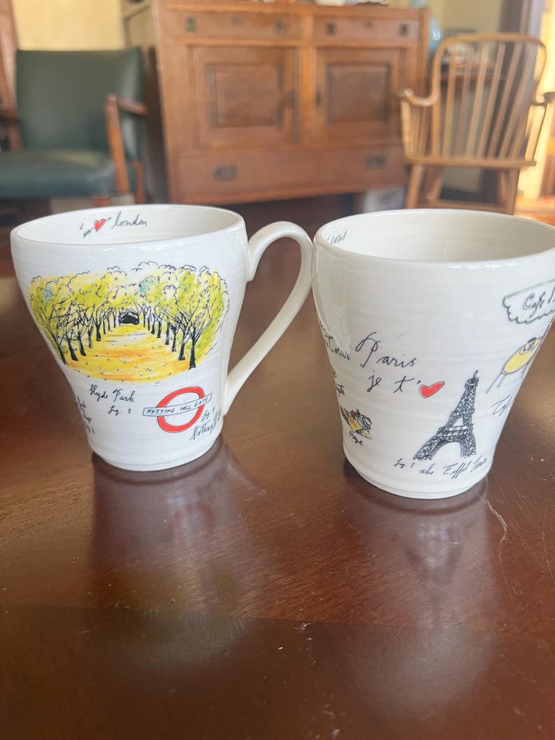 K&ouml;nnte beinhalten: Zwei wei&szlig;e Keramikbecher mit handgezeichneten Illustrationen. Ein Becher zeigt eine Londoner Szene mit dem Text "Hyde Park" und "Padding Hill Gate". Der andere Becher hat ein Pariser Thema mit einer Eiffelturm-Illustration und dem Text "Paris je t'aime".