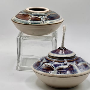 May include: Two ceramic oil lamps with a white glaze and blue and brown patterns. The lamp on the left is a simple bowl shape, while the lamp on the right has a round, slightly raised center and a wick.