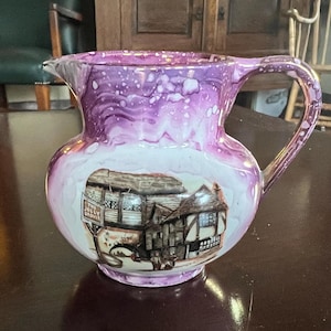 May include: A vintage ceramic jug with a rounded body and a handle. The jug has a white and purple marbled design with a detailed illustration of a building. The jug is likely for decorative use.