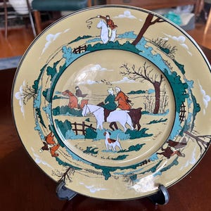 Puede incluir: Plato decorativo con una escena de caza. El plato presenta un fondo color crema con una ilustración detallada de jinetes a caballo, perros y árboles. El borde está acentuado con un ribete dorado y un borde verde y turquesa.