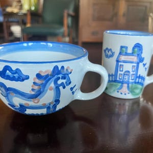 May include: Two white ceramic mugs with blue and pink painted designs. One mug is a cup with a horse design, and the other is a mug with a house design. Both mugs have blue rims and handles.