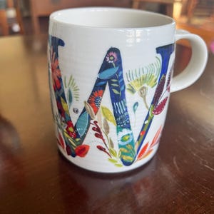 K&ouml;nnte beinhalten: Wei&szlig;e Keramiktasse mit einem gro&szlig;en, floralen "W"-Design. Der Buchstabe ist haupts&auml;chlich blau und mit bunten Blumen und Bl&auml;ttern gef&uuml;llt. Die Tasse hat einen gebogenen Henkel. Der Hintergrund ist unscharf.
