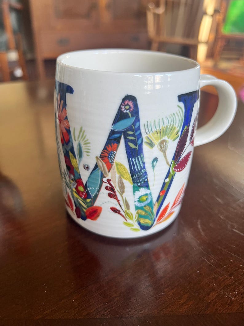 K&ouml;nnte beinhalten: Wei&szlig;e Keramiktasse mit einem gro&szlig;en, floralen "W"-Design. Der Buchstabe ist haupts&auml;chlich blau und mit bunten Blumen und Bl&auml;ttern gef&uuml;llt. Die Tasse hat einen gebogenen Henkel. Der Hintergrund ist unscharf.