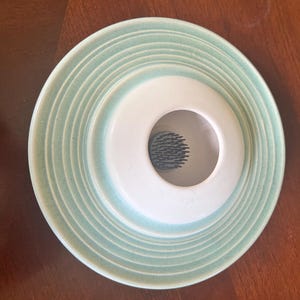 Può includere: Un ranocchio per fiori in ceramica con una combinazione di colori azzurro e bianco. Il ranocchio ha un design circolare con anelli concentrici e un'apertura centrale. L'interno dell'apertura presenta un motivo scuro.