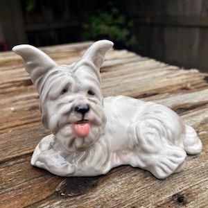 May include: A white ceramic figurine of a dog with its tongue sticking out. The dog is lying down with its head turned to the side. The figurine is detailed with realistic features, including fur and a pink tongue.