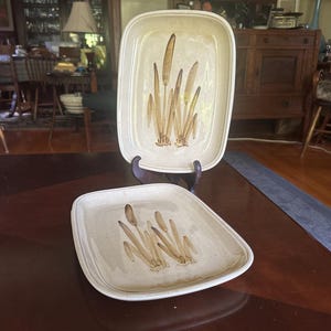 May include: Two rectangular ceramic serving platters with rounded corners, featuring a speckled cream-coloured glaze and a central brown botanical illustration of cattail plants. One platter is displayed upright on a stand, while the other lies flat.