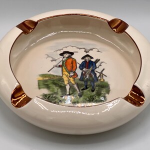 May include: Cream-colored ceramic ashtray with a brown rim and four cigarette rests. The interior features a painted scene of two figures in period clothing, set against a landscape with green grass and a blue sky.