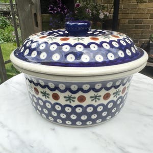 Puede incluir: Una fuente redonda de cerámica para horno con tapa a juego, que presenta una base de color crema decorada con un patrón repetitivo de círculos azules, pequeñas formas de estrella verdes y puntos naranjas. La tapa tiene un pomo azul oscuro.