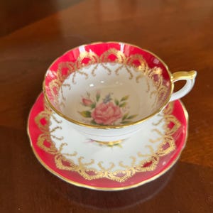 May include: A teacup and saucer set in red and gold with a floral design. The cup has a red rim and a gold trim. The saucer has a red rim and a gold trim. The floral design features a pink rose and green leaves.