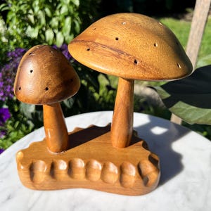 Könnte beinhalten: Eine Holzpilzskulptur mit zwei Pilzkappen und -stielen. Die Kappen haben kleine Löcher und sind an einer geschnitzten Basis befestigt. Die Skulptur ist aus hellbraunem Holz gefertigt und steht auf einer weißen Oberfläche.