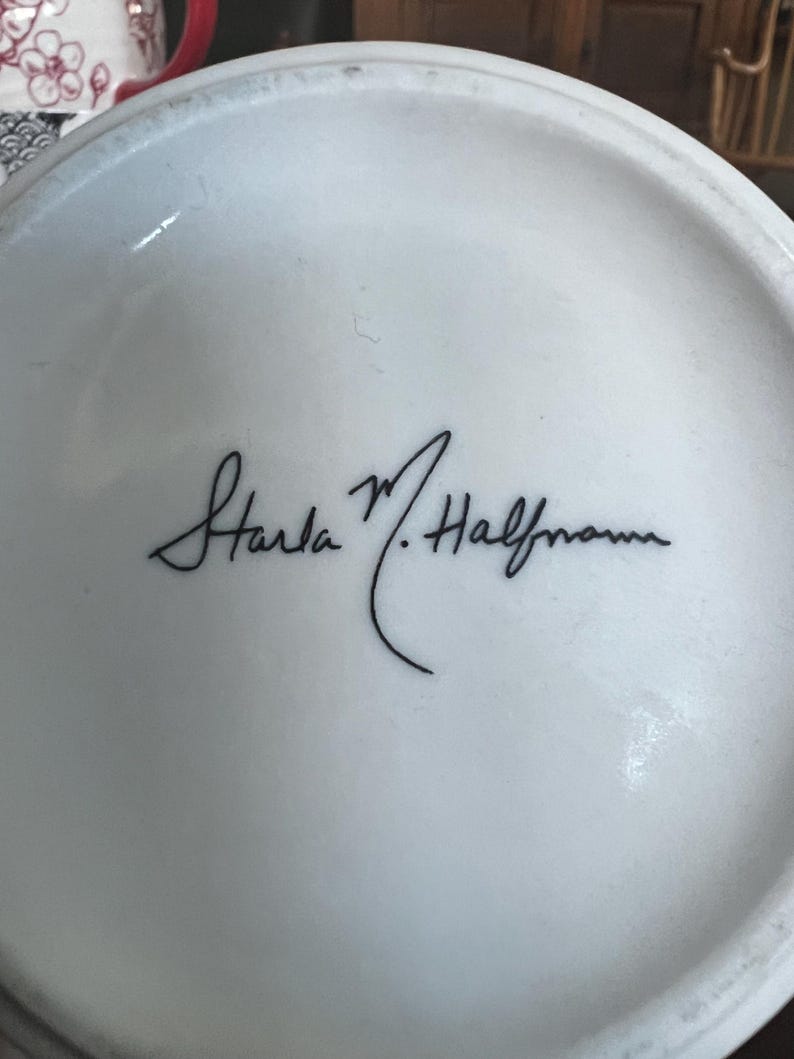 K&ouml;nnte beinhalten: Ein wei&szlig;es Keramikobjekt mit einer schwarzen Signatur, die "Starla M. Halfmann" lautet. Die Signatur ist in Schreibschrift. Das Objekt ist leicht gekr&uuml;mmt und scheint der Boden eines Keramikst&uuml;cks zu sein.