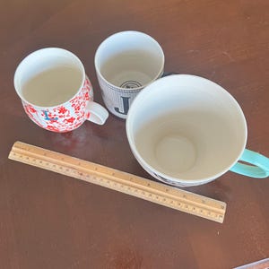 K&ouml;nnte beinhalten: Drei Keramikbecher in verschiedenen Designs und Gr&ouml;&szlig;en sind auf einer braunen Oberfl&auml;che angeordnet. Ein Becher hat ein rotes Blumenmuster, ein anderer ein schwarz-wei&szlig;es Muster mit dem Buchstaben "J", und der dritte ist haupts&auml;chlich wei&szlig; mit einem t&uuml;rkisfarbenen Griff.