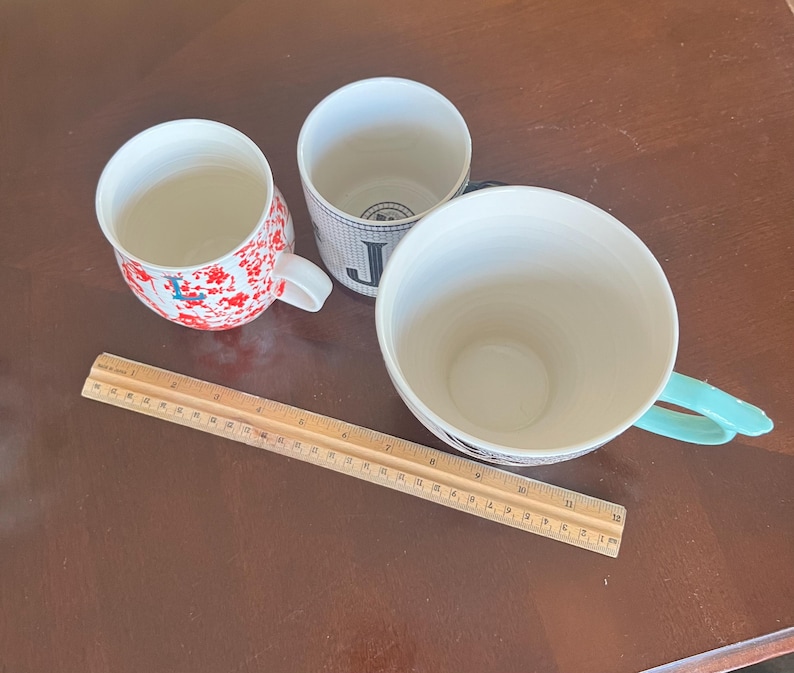 K&ouml;nnte beinhalten: Drei Keramikbecher in verschiedenen Designs und Gr&ouml;&szlig;en sind auf einer braunen Oberfl&auml;che angeordnet. Ein Becher hat ein rotes Blumenmuster, ein anderer ein schwarz-wei&szlig;es Muster mit dem Buchstaben "J", und der dritte ist haupts&auml;chlich wei&szlig; mit einem t&uuml;rkisfarbenen Griff.
