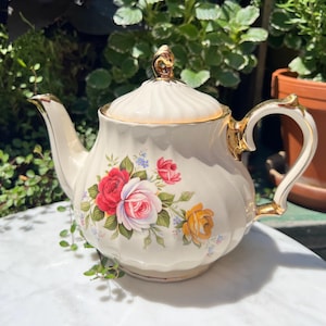 May include: Cream-colored porcelain teapot with a lid, adorned with a floral rose design in red, pink, and yellow. The teapot has gold trim on the handle, spout, and lid finial. A classic tea set item.