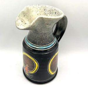 Peut inclure: Un petit pichet en céramique noire avec un glaçage blanc moucheté sur le dessus. Le pichet a un bec unique et incurvé et une anse. Il est décoré d'un motif géométrique bleu, jaune et orange.