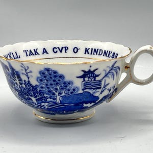 Puede incluir: Taza de té blanca con un diseño de paisaje azul y blanco y ribete dorado. El borde de la taza tiene el texto azul "WELL TAK A CUP O' KINDNESS". La taza tiene un asa y un pie pequeño.