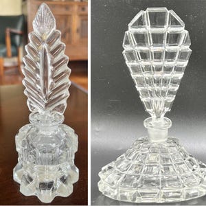 Puede incluir: Dos frascos de perfume de vidrio transparente. Uno tiene un tapón en forma de hoja y el otro un diseño geométrico. Ambos frascos están vacíos y parecen ser antiguos. Los frascos se exhiben sobre una mesa y una superficie oscura.
