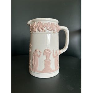 May include: A white ceramic pitcher with a pink relief design. The pitcher features a handle and a spout. The relief design includes grape vines and classical figures. The pitcher is likely for decorative use.