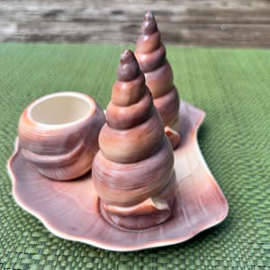 Puede incluir: Un juego de cerámica con temática de conchas marinas, que presenta dos candelabros en forma de espiral y una taza pequeña, todo sobre una bandeja a juego con forma de concha. El juego está pintado en tonos marrones y rosas, creando una estética cálida y natural.