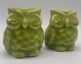 May include: Two green ceramic owl figurines. The owls have a glazed finish and are sitting on a white surface.