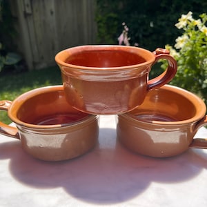 Puede incluir: Tres tazas de cerámica marrón con asas redondeadas. Las tazas están apiladas una encima de la otra sobre una superficie blanca.