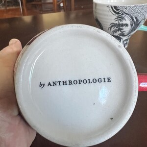 K&ouml;nnte beinhalten: Wei&szlig;er Keramik-Tassenboden mit dem Text "by ANTHROPOLOGIE" in Schwarz. Die Tasse steht auf einer dunkelbraunen Oberfl&auml;che. Eine weitere Tasse mit einem schwarz-wei&szlig;en Design befindet sich im Hintergrund.