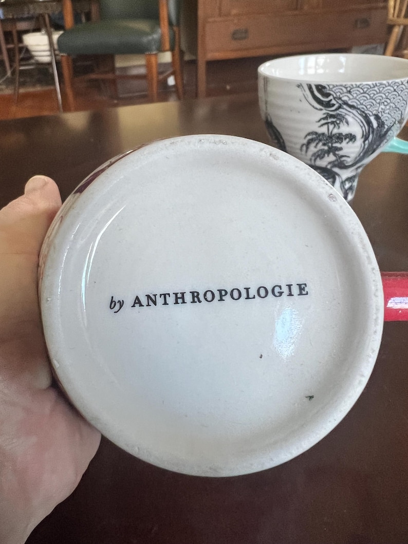 K&ouml;nnte beinhalten: Wei&szlig;er Keramik-Tassenboden mit dem Text "by ANTHROPOLOGIE" in Schwarz. Die Tasse steht auf einer dunkelbraunen Oberfl&auml;che. Eine weitere Tasse mit einem schwarz-wei&szlig;en Design befindet sich im Hintergrund.