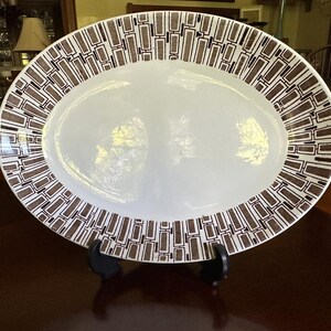 May include: An oval ceramic serving platter with a white centre and a wide border featuring a repeating geometric pattern of brown rectangular shapes and thin black lines. The platter is displayed upright on a black plastic stand.