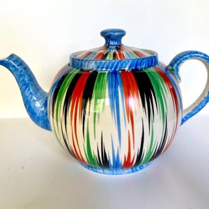 Puede incluir: Una tetera de cerámica con cuerpo redondo y tapa. La tetera está decorada con rayas verticales en rojo, negro, verde, azul y blanco. El asa y el pico son azules.