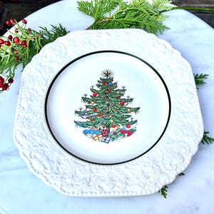 May include: White ceramic Christmas plate with an embossed border. The plate features a central image of a decorated Christmas tree with ornaments and wrapped gifts. A black rim surrounds the tree image.