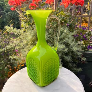 Op de afbeelding: Een felgroene glazen kan met een getextureerd ontwerp en een heldere glazen handvat. De kan staat op een wit marmeren oppervlak.