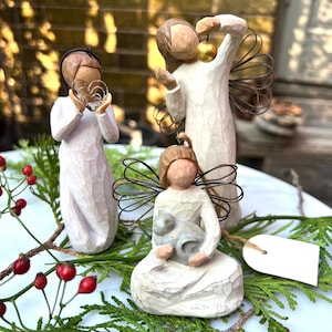 Puede incluir: Tres figuras de ángeles con túnicas blancas y alas de alambre están dispuestas sobre una superficie blanca con vegetación y bayas rojas. Un ángel sostiene un corazón, otro un adorno dorado y el tercero acuna un cordero. Las figuras tienen un aspecto rústico tallado.