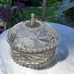 Pode incluir: Uma bomboniere de vidro transparente com base e tampa de metal prateado. A tampa tem um botão prateado e é decorada com padrões florais. A base tem um design cruzado. O prato está sobre uma mesa de mármore branco.