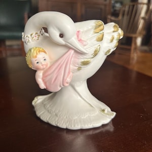 Puede incluir: Una figura de cerámica blanca de una cigüeña que lleva un bebé en una manta rosa. La cigüeña tiene detalles dorados en sus alas y un diseño floral alrededor de su cuello. El bebé tiene el pelo rubio.