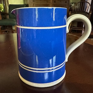 Peut inclure: Un pichet cylindrique en céramique arborant un glaçage bleu cobalt vif avec deux bandes horizontales blanches distinctes entourant le corps. Le récipient comprend une anse blanche incurvée sur le côté et un petit bec verseur au sommet.