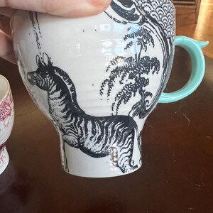 K&ouml;nnte beinhalten: Wei&szlig;er Keramikbecher mit einer schwarzen Zebra-Illustration und einem hellblauen Griff. Der Becher zeigt ein detailliertes schwarz-wei&szlig;es Design eines Zebras, B&auml;umen und anderen dekorativen Elementen.