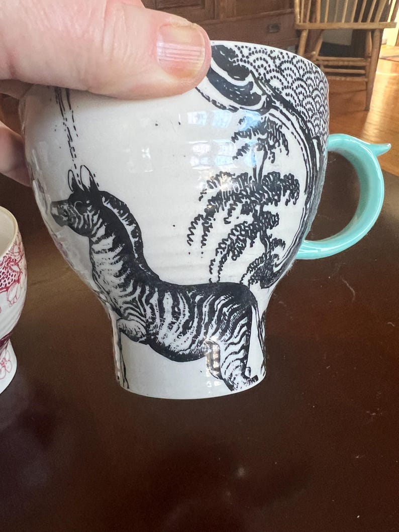 K&ouml;nnte beinhalten: Wei&szlig;er Keramikbecher mit einer schwarzen Zebra-Illustration und einem hellblauen Griff. Der Becher zeigt ein detailliertes schwarz-wei&szlig;es Design eines Zebras, B&auml;umen und anderen dekorativen Elementen.