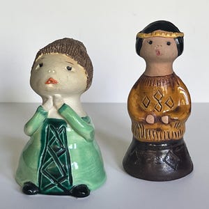 May include: Two ceramic figurines. One is a woman in a green dress with a dark green design. The other is a Native American figure in a yellow and brown top with a black base. Both are vintage and decorative.