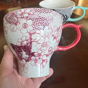K&ouml;nnte beinhalten: Wei&szlig;e Keramiktasse mit rotem Blumen- und Katzenmuster. Die Tasse hat einen roten Henkel. Eine weitere Tasse mit blauem Henkel und einem schwarz-wei&szlig;en Muster befindet sich im Hintergrund. Die Tasse wird in einer Hand gehalten.