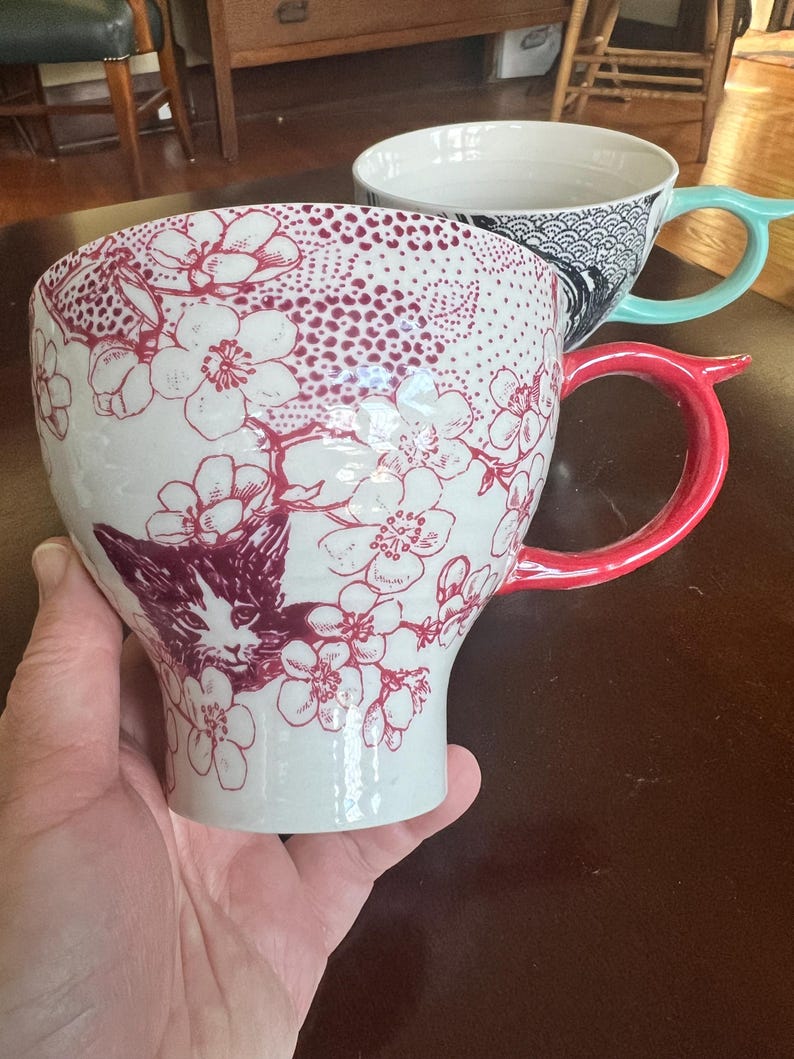 K&ouml;nnte beinhalten: Wei&szlig;e Keramiktasse mit rotem Blumen- und Katzenmuster. Die Tasse hat einen roten Henkel. Eine weitere Tasse mit blauem Henkel und einem schwarz-wei&szlig;en Muster befindet sich im Hintergrund. Die Tasse wird in einer Hand gehalten.