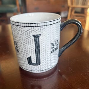 K&ouml;nnte beinhalten: Wei&szlig;e Keramiktasse mit schwarzem Henkel und schwarzem Buchstaben "J" auf der Vorderseite. Die Tasse hat ein Wabenmuster und florale Akzente. Die Tasse steht auf einer dunkelbraunen Holzoberfl&auml;che.