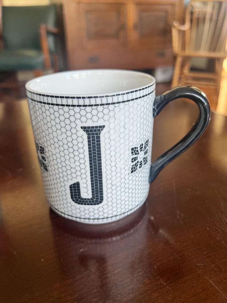 K&ouml;nnte beinhalten: Wei&szlig;e Keramiktasse mit schwarzem Henkel und schwarzem Buchstaben "J" auf der Vorderseite. Die Tasse hat ein Wabenmuster und florale Akzente. Die Tasse steht auf einer dunkelbraunen Holzoberfl&auml;che.